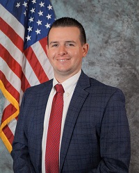 Picture of Detective Sergeant Lee olds dressed in business attire, from approximately waist up, sitting in front of an American flag.